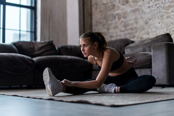 Slim sportswoman in sportswear warming-up muscles before workout doing stretching exercise sitting on floor at home - Powered by Adobe