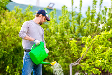 Smiling farmer in cap is pouring water from watering can at spring garden background. © Mayatnikstudio
