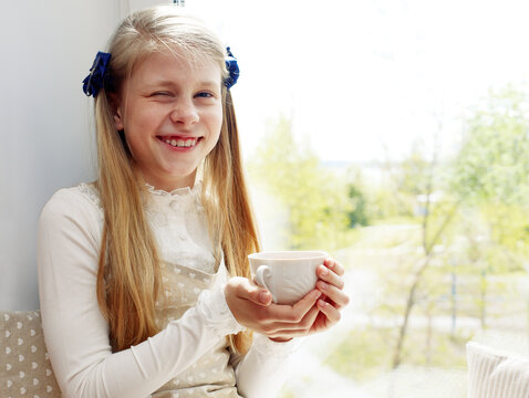 Beautiful child drinking tea and looking through window while sitting at windowsill at home