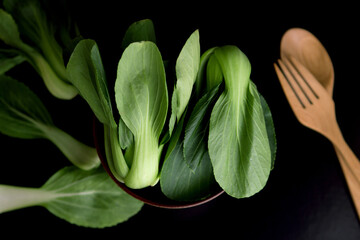 close up Fresh baby green bok choy in bowl with woodden spoon and fork on the  black background , overhead or top view shot - obrazy, fototapety, plakaty