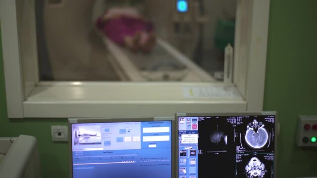 Crane motion, tilt down from patient behind protective glass who lying on CT or MRI scanner blurred to monitor screen with x-ray images of human body that changes in focus, shallow depth of field