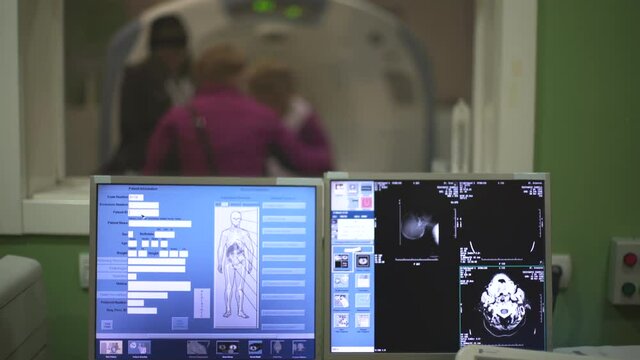 People help elderly female patient to stand up from CT or MRI scanner after scanning blurred, crane shot, tilt down on x-ray images on monitor screen in focus, shallow depth of field, room interior.