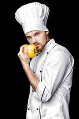 Young bearded man chef In white uniform holds yellow bell pepper on  black background