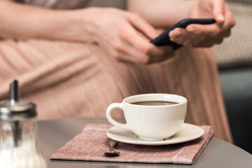 Close-up partial view of woman using smartphone and white cup with coffee on table