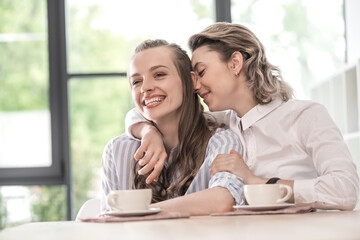 young smiling caucasian girlfriends drinking coffee and embracing at cafe