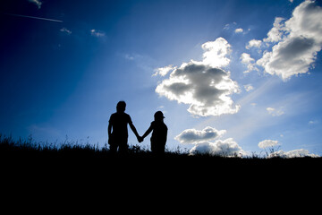 Fototapeta premium The silhouettes of two children holding hands on the top one hill and the clear sky with some clouds