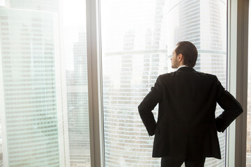 Fototapeta premium Back view of man in suit standing with hands on hips looking on cityscape outside the window. Successful businessman thinking about future plans, guy dreams about great career. Ambitions in business