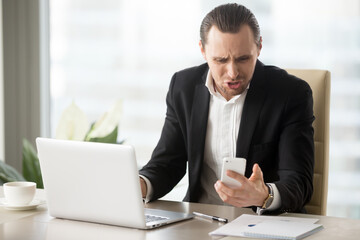 Fototapeta na wymiar Businessman angry because of ill-timed or annoying phone call in time his working on laptop at office. Entrepreneur annoyed with cellphone breakdown or mobile app critical error, bad news in message
