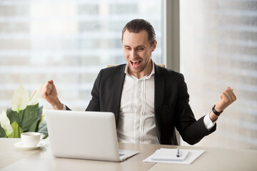 Fototapeta premium Happy businessman excited because of great deal, achievement, company success. Young entrepreneur screaming with joy while looking on laptop screen. Man overflowing with positive emotions at workplace