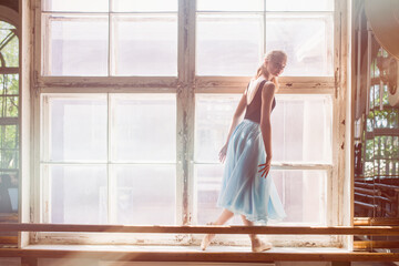 A ballerina is posing in front of a large window, standing on the windowsill. Girl in a turquoise ballet skirt.