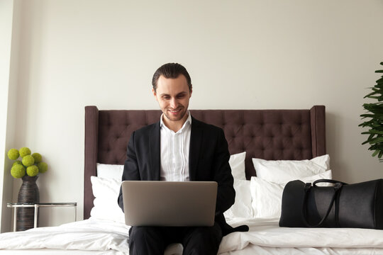 Happy man wearing business suit sitting on bed with luggage and working on laptop. Businessman ready to travel orders airline tickets online. Entrepreneur searching in Internet vacation travel tour