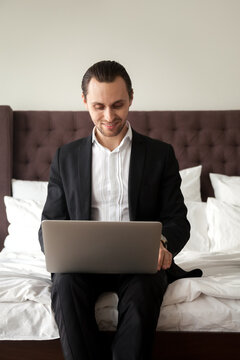 Happy businessman working on laptop while sitting on bed in bedroom. Business traveler finalize project on computer in hotel room. Entrepreneur checks e-mail, reading news at home before going to work