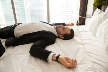 Young man in business suit lying with arms outstretched on bed with white sheets. Businessman resting at home after hard work day. Entrepreneur sleeps in comfortable hotel room after long air flight