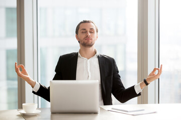 Relaxed young entrepreneur meditating at desk in front of laptop. Office worker doing yoga...