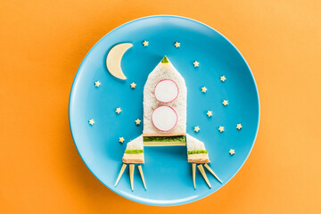 top view of funny colorful breakfast for child in shape of rocket with moon and stars on blue plate isolated on yellow