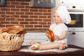 Little smiling baby girl baker in white cook hat and apron kneads a dough and holds a bagel