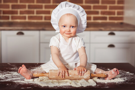 Little smiling baby girl baker in white cook hat and apron kneads a dough at brick wall background