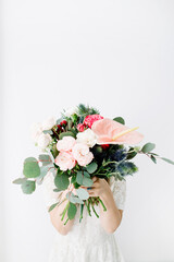 Pretty woman with beautiful flowers bouquet: bombastic roses, blue eringium, anthurium flower, eucalyptus branches at white wall. Front view. Floral lifestyle composition.