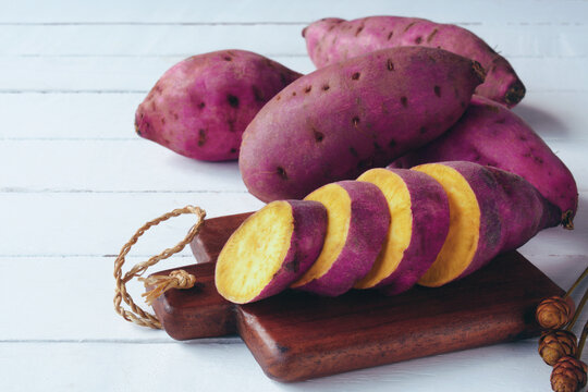 raw sweet potatoes sliced on chopping board with potato root in purple skin on white wooden background