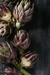 Fototapeta premium Uncooked whole and sliced organic wet purple artichokes with salt over dark wooden background. Rustic style. Top view with space