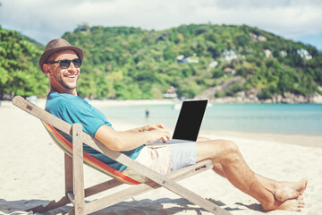 Attractive young man with laptop working on the beach. Freedom, remote work, freelancer,...