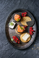 Stack of black charcoal and traditional crackers with smoked salmon, cream cheese, green salad and red currant berries on vintage metal tray over black stone background. Appetizer snack. Top view