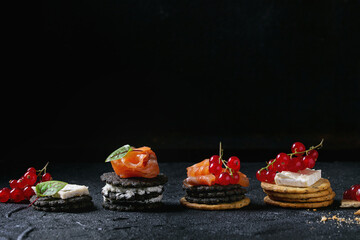 Stack of black wholegrain charcoal and traditional crackers with smoked salmon, cream cheese, green salad and red currant berries over black stone background. Appetizer snack. Space for text