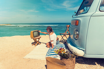 Woman is sitting on the beach for a picnic near a vintage car, next to an old TV set. Summer holidays, road trip, vacation, travel and people concept - smiling young women next a vintage minivan car