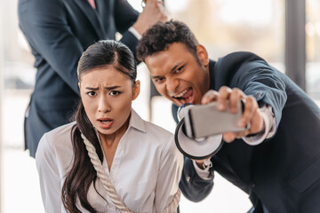 asian businesswoman bound with rope while businessman taking selfie and screaming in megaphone