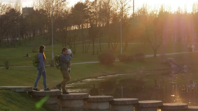 Active family with the baby passes a beautiful bridge over the river at sunset. The sun shines in the frame