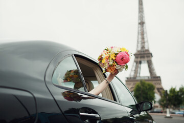 Bride reaches out hand with wedding bouquet from a car before Eiffel Tower
