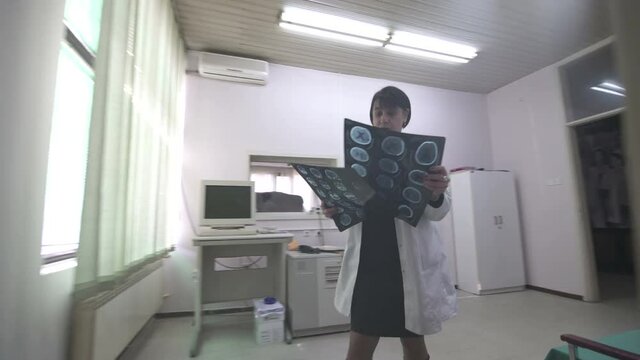 Middle aged female doctor specialist in white gown comes in consulting room, puts x-rays on the light board and sits at computer to examine images, steady cam tracking shot, active real scene.
