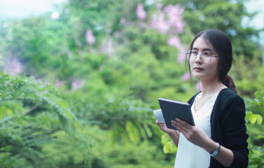 Fototapeta premium Happy young student with a tablet and a disposable coffee cup sitting on the bench and reading in a summer park.