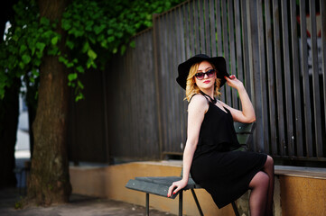 Blonde woman on black dress, necklaces and hat sitting on bench.