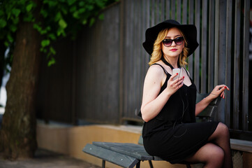 Obraz premium Blonde woman on black dress, sunglasses, necklaces and hat sitting on bench with mobile phone at hand and headphones.