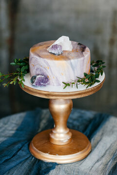 unusual weeding cake decorated like marble with green brunches and white and purple quartz stones on wooden stand. Chic vintage boho style