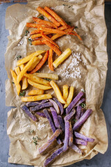 Variety of french fries traditional potatoes, purple potato, carrot served with salt, thyme on baking paper over gray metal background. Top view with space. Homemade fast food