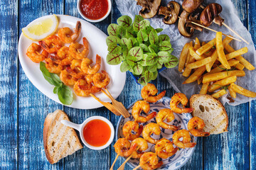 Variety of BBQ snack lunch. Plates grilled spicy king prawn kebabs, mushrooms skewers, bread, french fries potatoes with sauces and greens over blue wooden background. Flat lay