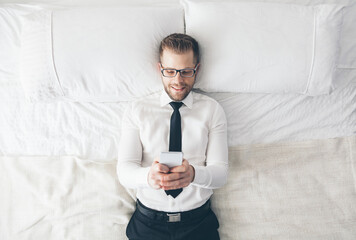 Top view. Handsome businessman with glasses lying on bed texting from his smartphone