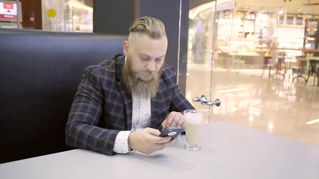Fashionable young man in plaid suit with a stylish haircut and a big beard sitting at a cafe table with a glass of iced coffee and writes the message on the mobile phone.