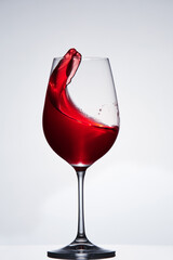 Splashing and moving red wine in the pure wineglass standing against light background with reflection on the stand.