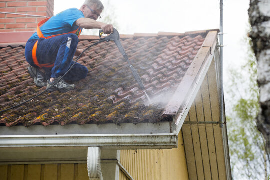 Caucasian man is washing the roof with a high pressure washer. He is wearing safety harness on a slippery roof.