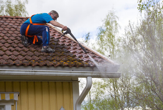Caucasian man is washing the roof with a high pressure washer. He is wearing safety harness on a slippery roof.