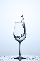 Trasparent water splash out of wineglass while standing on the glass with droplets against light background.