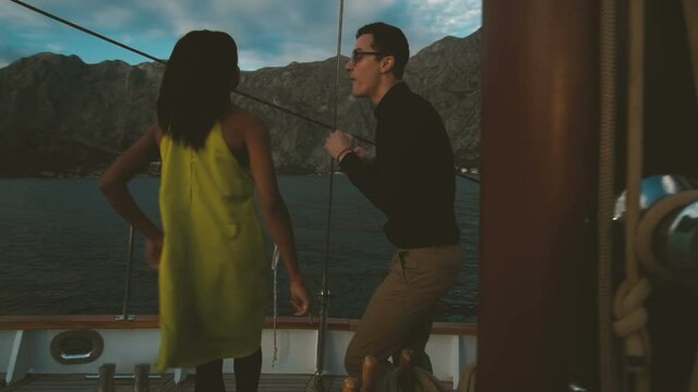 Romantic traveling of loving couple dancing on yacht outdoors. Beautiful woman with dark skinned hair yellow dress smiling dance enjoy music. man in shirt trousers looks at wife moving body. expensive