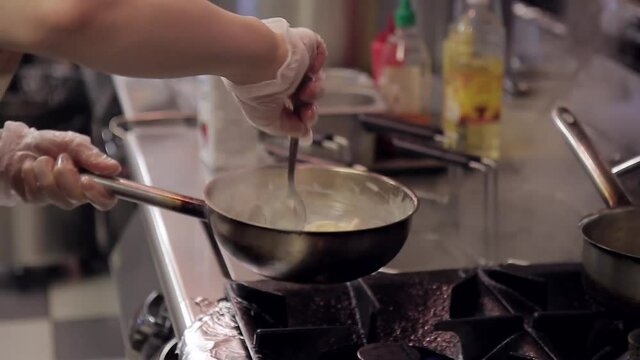 The chef prepares spaghetti on a gas flame in the restaurant kitchen. The cook in disposable gloves stirs pasta and sauce from milk with metal spoon. Man holds in his hand stewpan over black gas stove