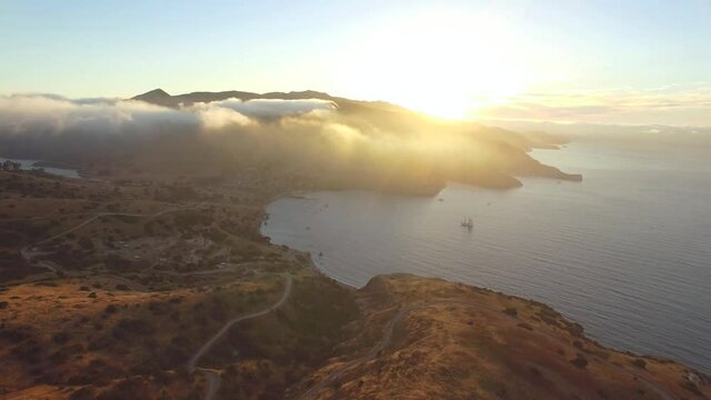 Aerial shot flying forwards towards a town, overlooking the coastline of an island at sunset. Gorgeous dazzling sunlight and cloud bank. A road winds along the cliff. sun reflects off water surface.