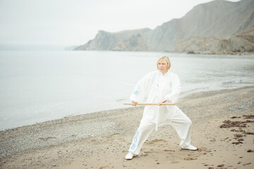 European caucasian adult woman In white clothes practicing Chinese traditional art Taijiquan in beautiful nature. Exercise with staff in morning day on the sea coast. Selective focus 