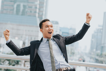 Celebrating success. Excited young Asian businessman arms up while standing outdoors with city  in ...