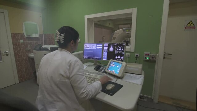 Steady cam shot from the body images on the computer screen to the female radiology technician sits at the table and scan male patient that lying on the MRI scanner behind protective glass, indoors.